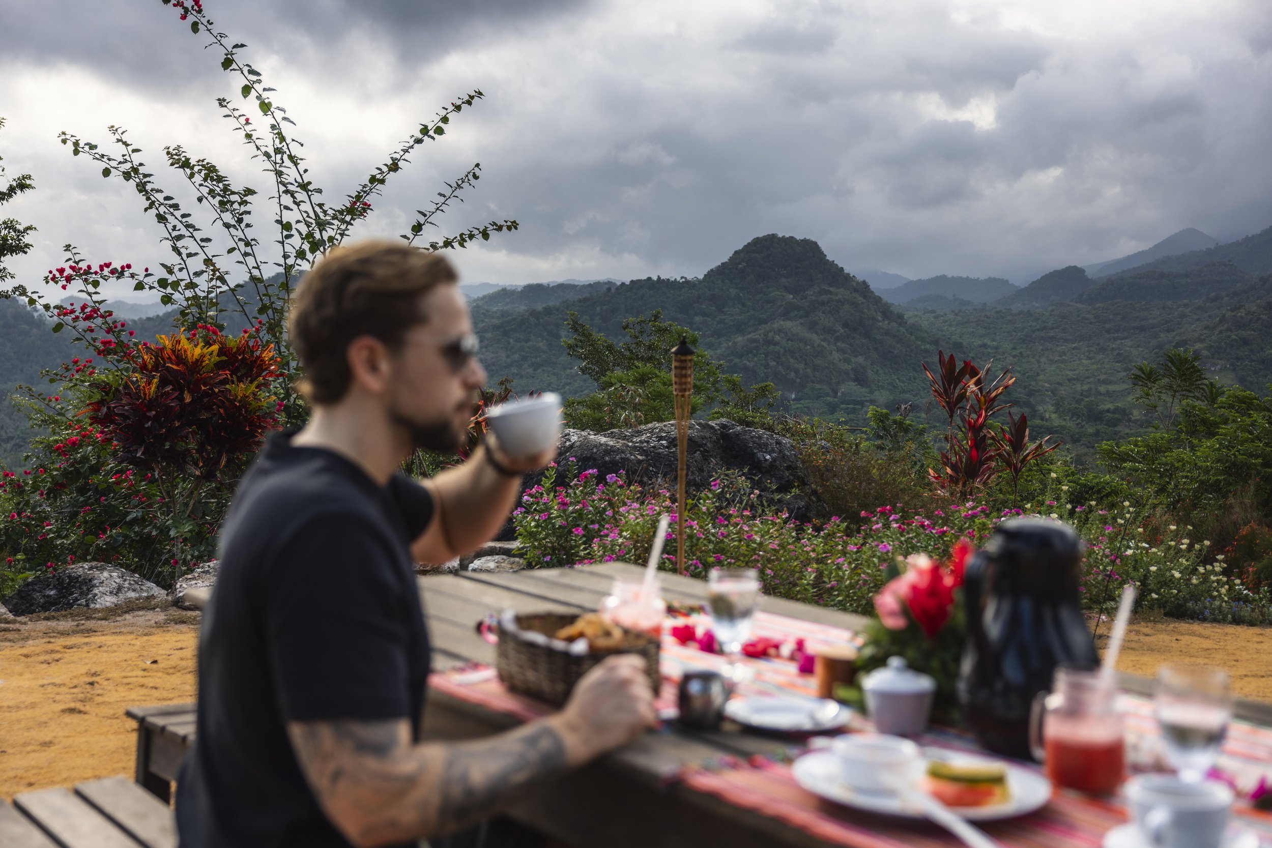 A person with short brown hair, glasses, and tattoos on his arms, sitting at an outdoor table with food and drinks, overlooking lush green mountains and cloudy skies.