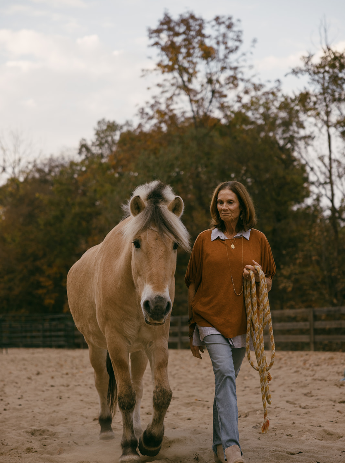 A woman walking with a light-colored horse in an outdoor equestrian arena during fall with trees in the background.