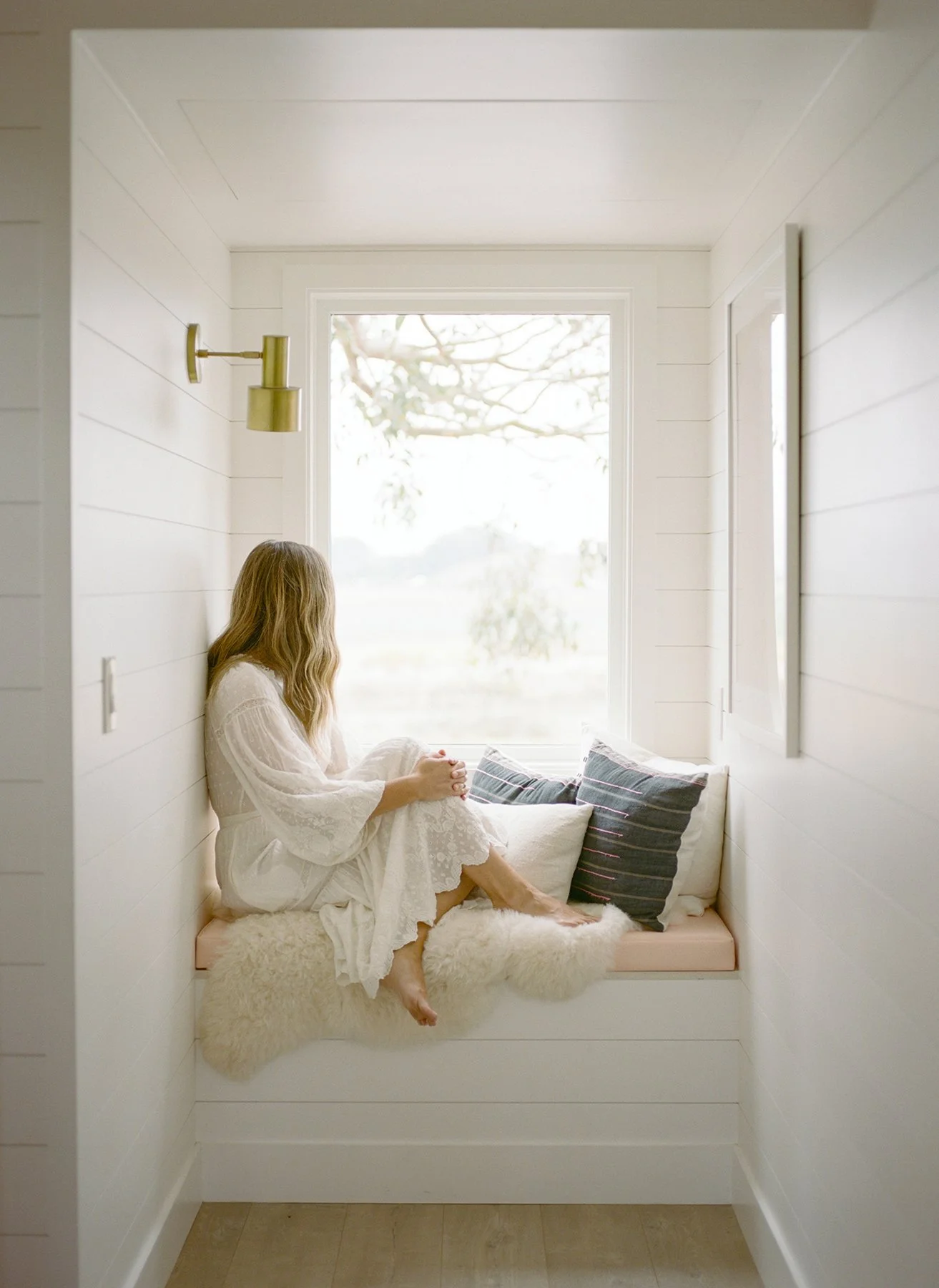 A woman sitting on a built-in window seat in a white-walled room, gazing out the window with a landscape view of trees and a cloudy sky.