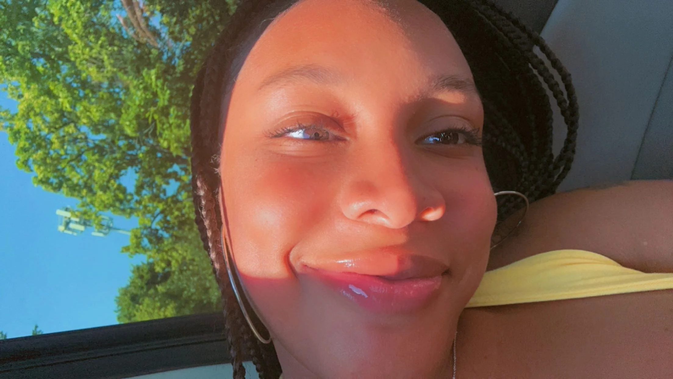 Close-up of a young woman with braided hair, smiling, with sunlight on her face, and trees visible in the background.