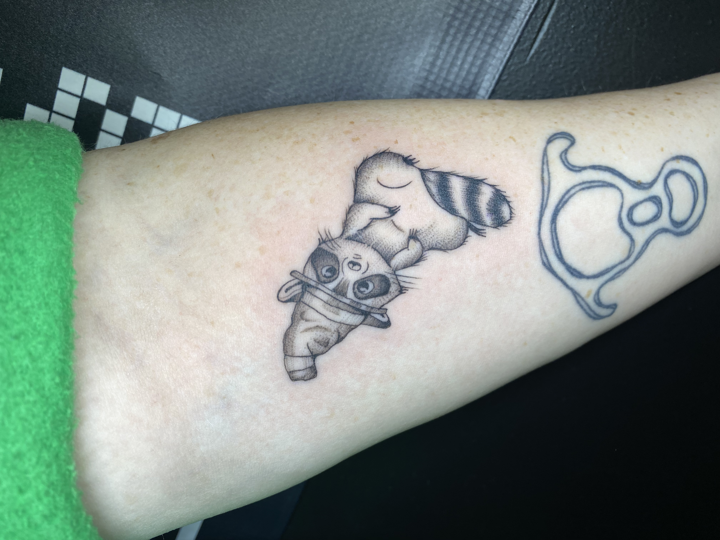 Tattoo of a cartoon animal, possibly a raccoon, holding a leaf to its mouth on a person's arm, with a separate outline tattoo of a ring or circular object nearby.