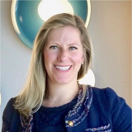 A woman with blonde hair smiling in front of a beige wall with a circular mirror.