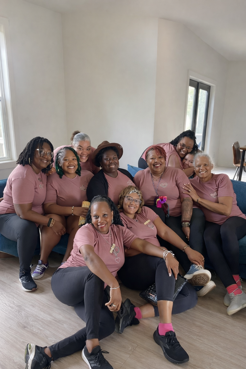 A group of ten women wearing matching pink T-shirts sitting on a blue sofa in a bright room with white walls and windows, smiling and posing for a photo.