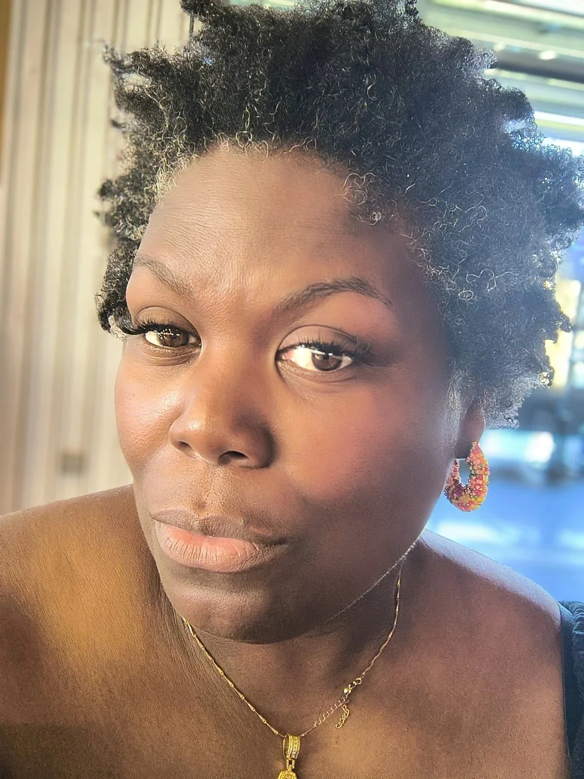 A close-up portrait of a woman with short, curly black hair, wearing colorful earrings and a gold necklace, sitting indoors near a window with daylight shining in.
