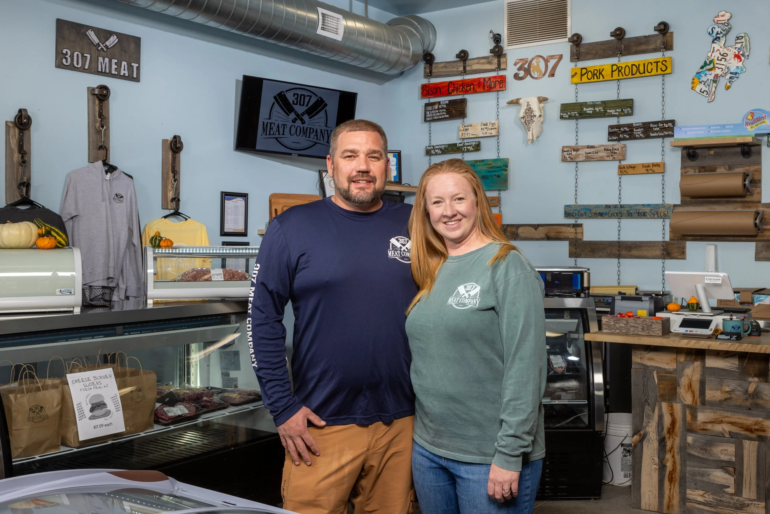 Business group portrait including owners of a business in Laramie, Wyoming including two people standing inside a meat shop, wearing shirts with the 307 Meat Company logo, smiling at the camera.