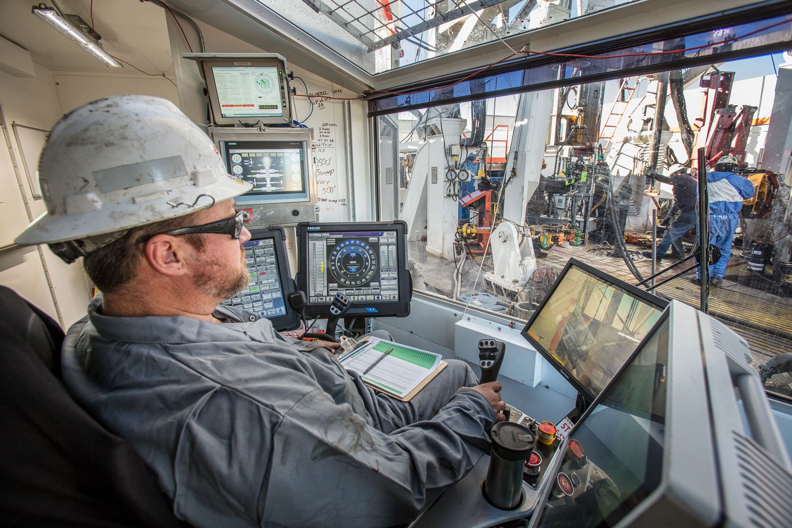 Drilling rig operator inside a drilling rig cabin operating controls while observing workers outside at a drilling deck near Parachute, Colorado.