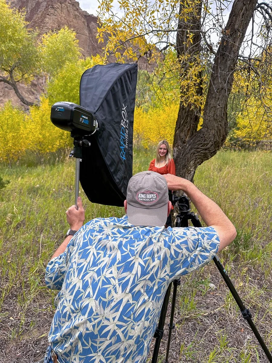 A professional photographer taking a portrait of a smiling young woman outdoors in a natural setting with trees and rocks, using studio lighting equipment and a camera on a tripod.