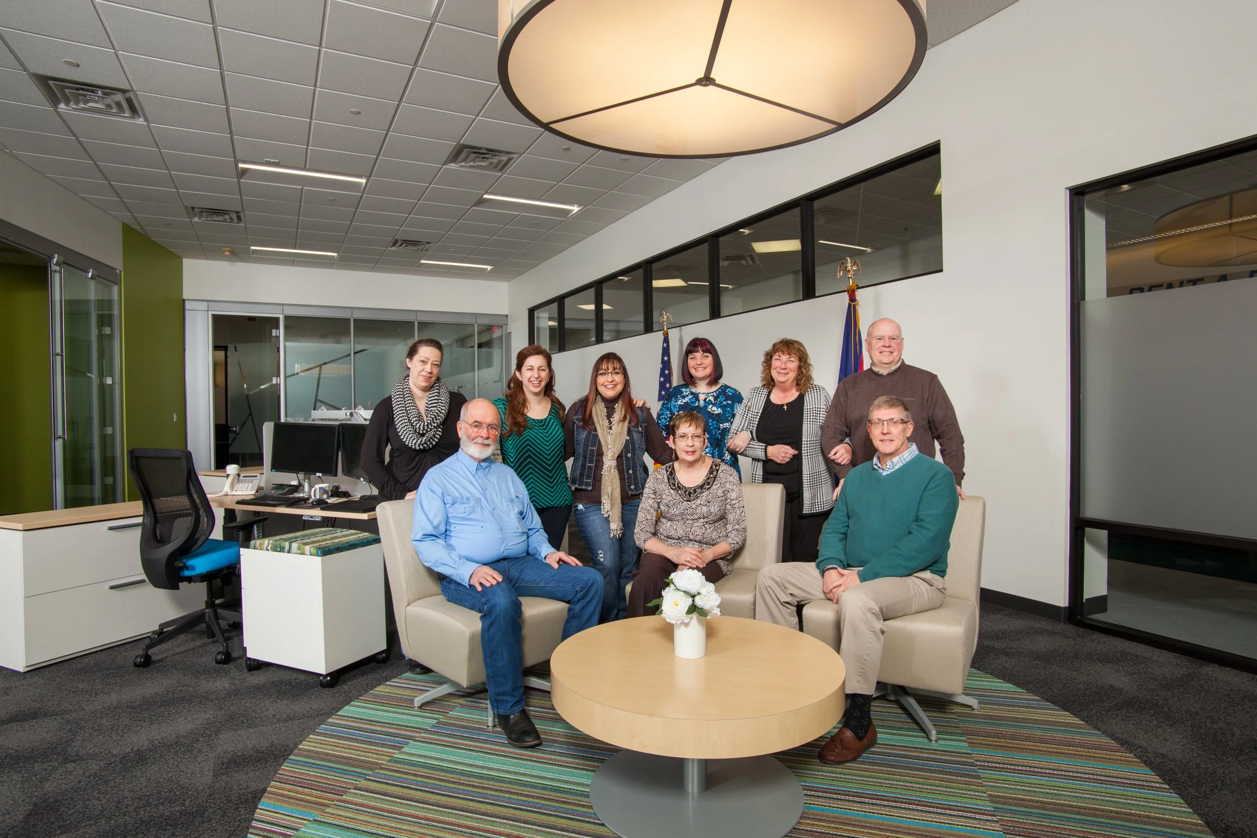 A business group portrait of nine people posing in a modern office lounge with a coffee table, sofa, and chairs. The background features office desks, a glass partition, and American flags.