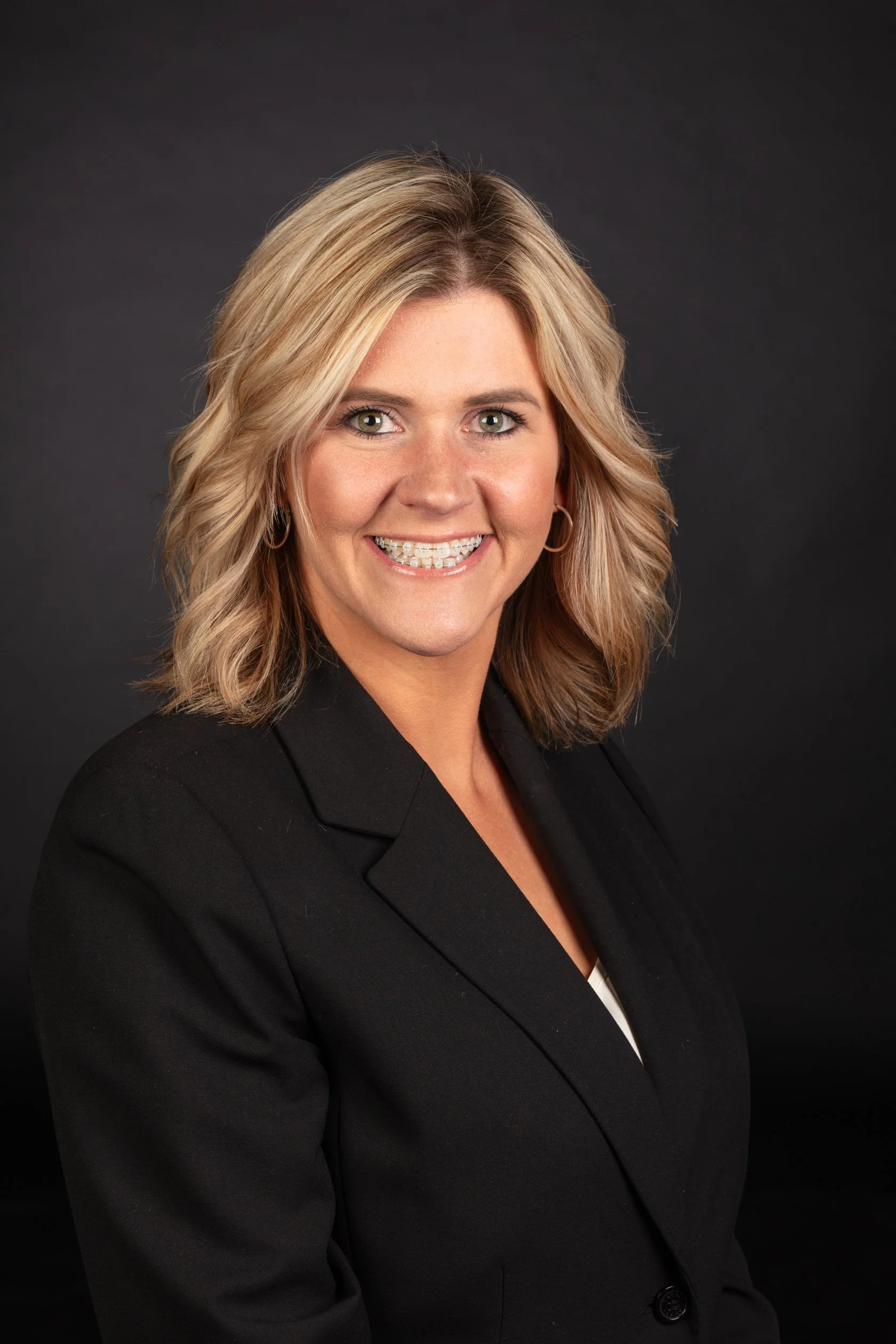 Business headshot portrait for marketing in Casper, Wyoming, including a woman with blonde hair wearing a black blazer and smiling against a dark background.