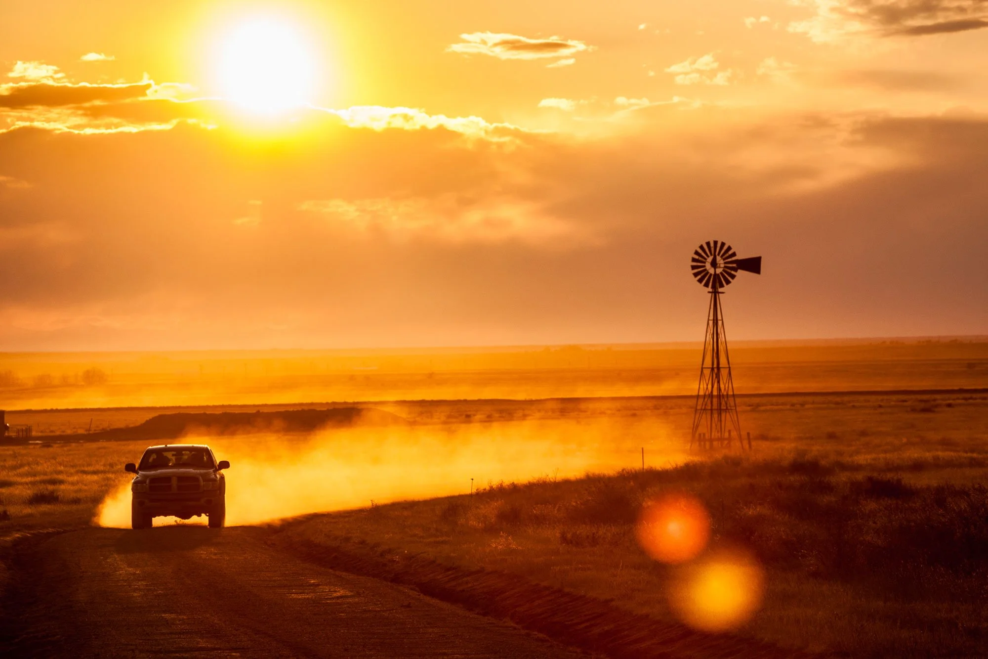 Ranch pickup travels down a dirt road after checking water at a windmill for his cattle at sunrise with dirt blowing up from the road.