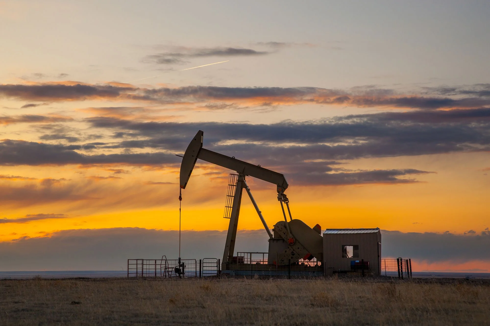 Silhouette of an oil pumpjack at sunset with a fence in the foreground and a cloudy sky in the background in Northeastern Colorado.