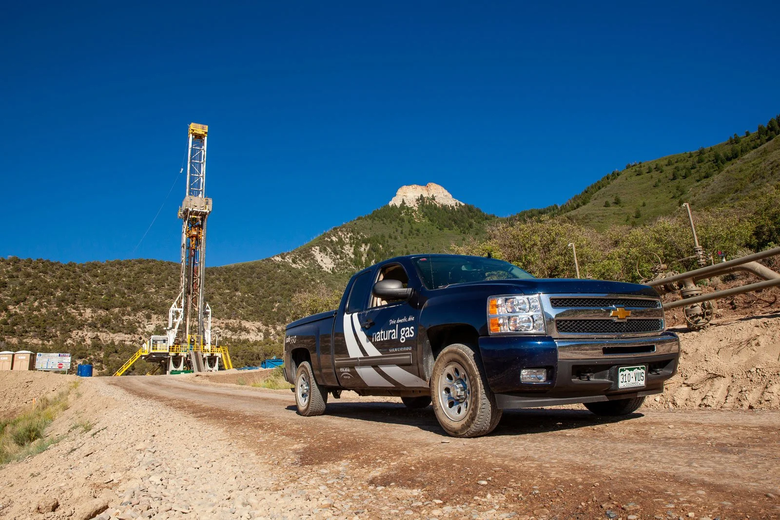 A black Chevrolet Silverado pickup truck traveling down a dirt road near an oil drilling rig, with mountains and a clear blue sky in the background near Parachute, Colorado.