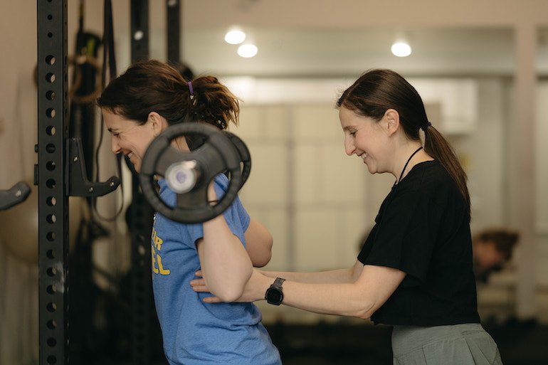 A woman helping a beginner strength trainer lift a barbell in a Toronto, inclusive gym, both smiling, with gym equipment in the background.
