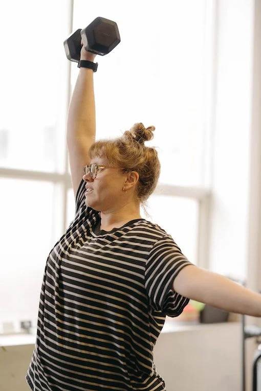 A woman wearing glasses and a striped t-shirt lifting a dumbbell in neighbourhood gym in Toronto.