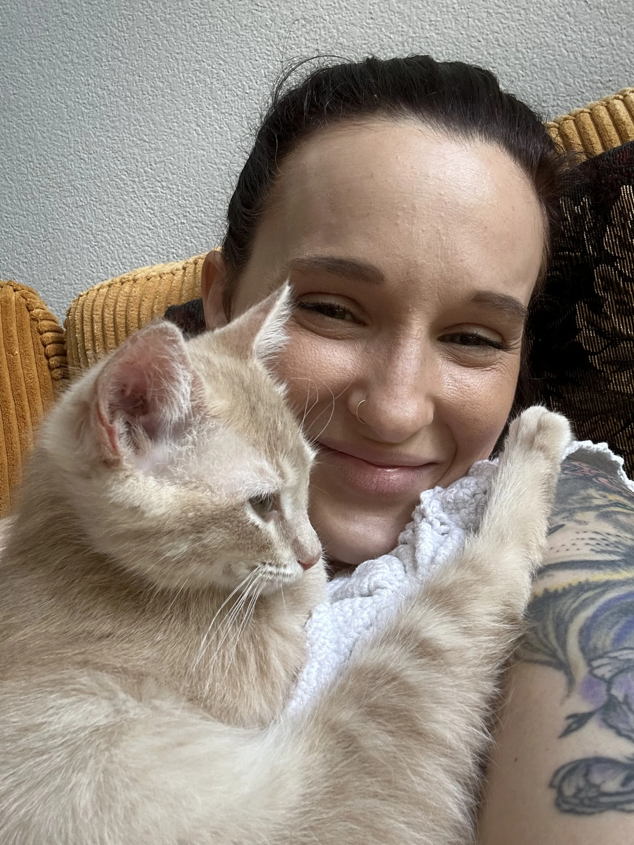 A woman with dark hair pulled back, smiling with a nose ring, hugging a light-colored cat with tabby markings.