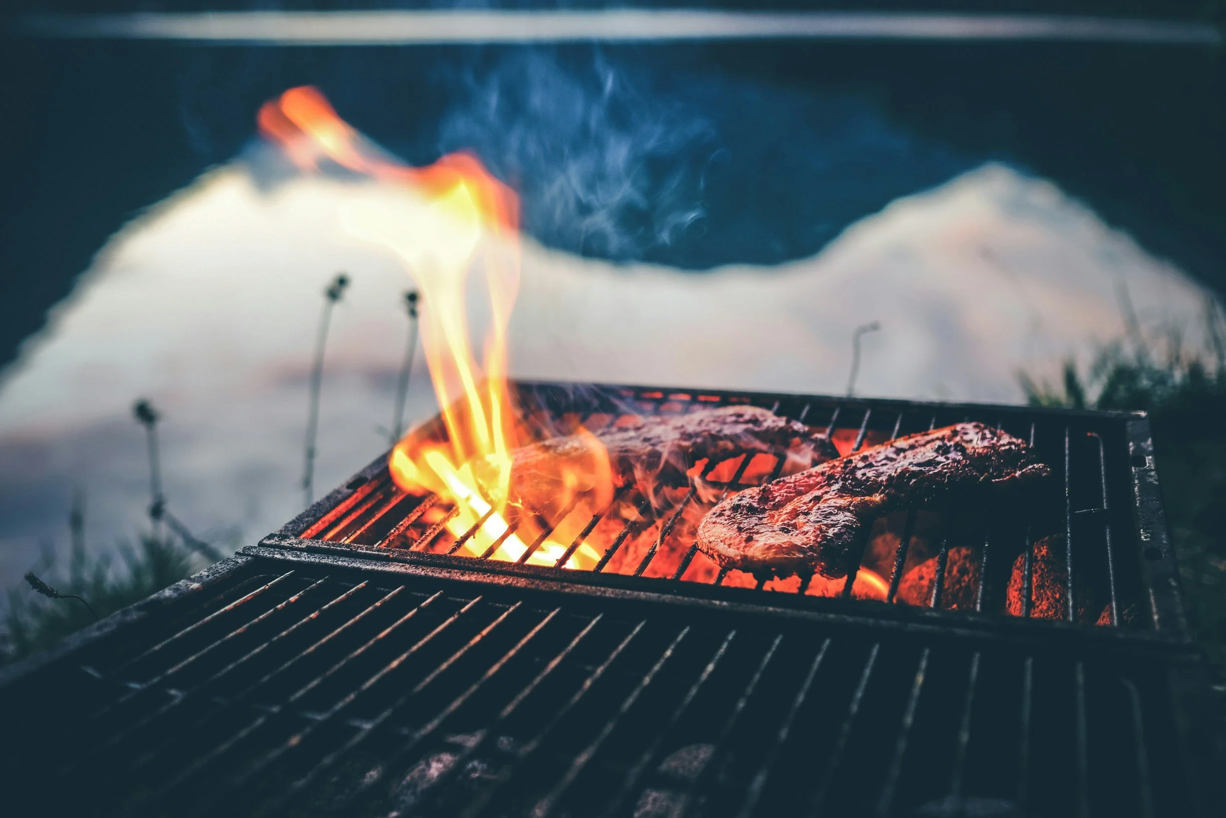 Meat cooking on a charcoal grill with flames and smoke rising from the grill.