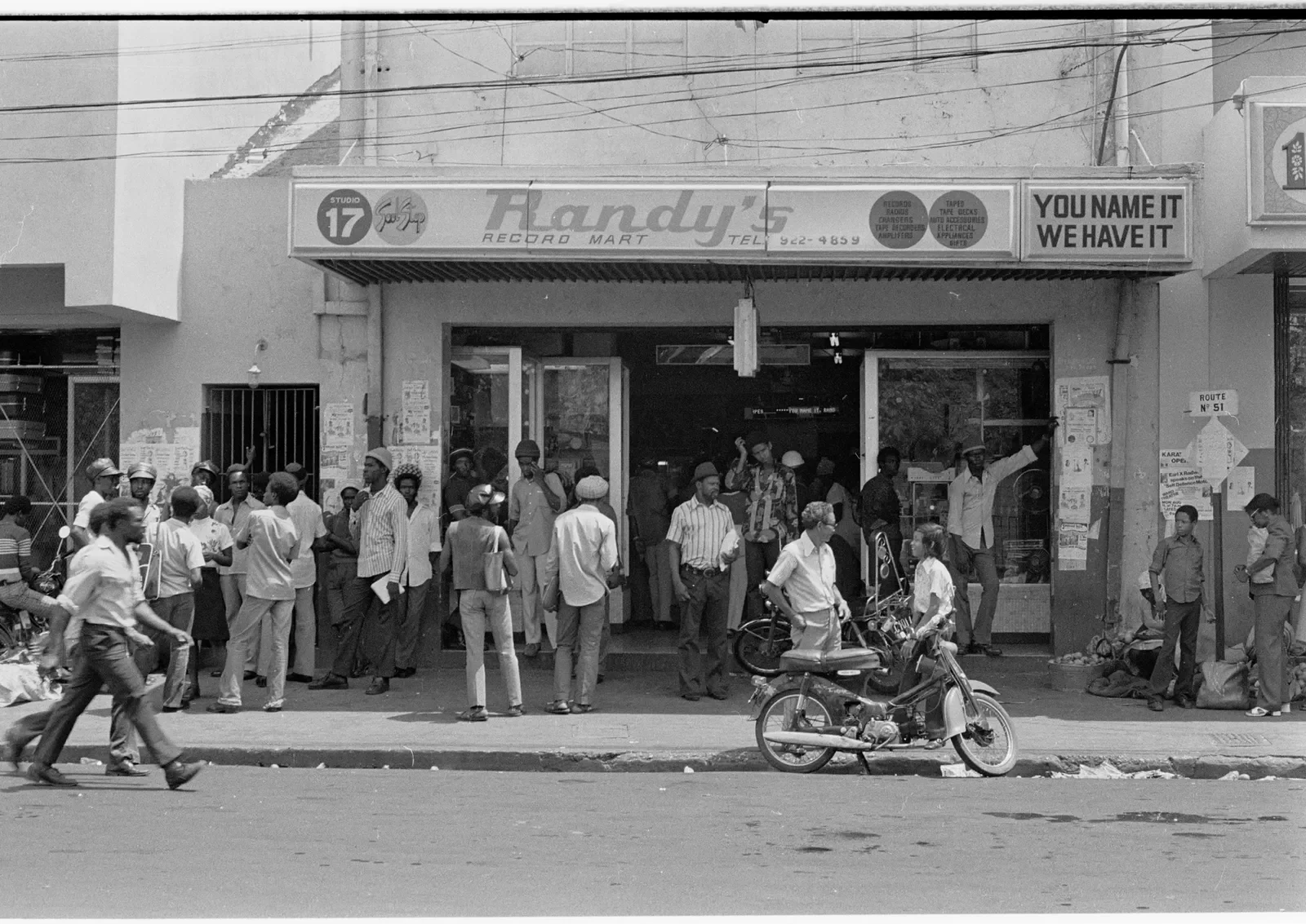 Street scene in front of a store with people standing and walking outside, some wearing helmets, and a scooter parked on the sidewalk.