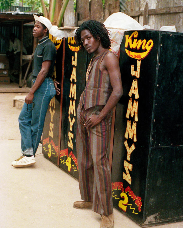 Two young men standing outdoors next to a large black vending machine with colorful writing that says 'King Sammy's' and 'Super Power.' One man with shoulder-length dreadlocks wears striped pants and a sleeveless shirt, leaning against the vending machine, while the other in a cap, short-sleeve shirt, and jeans stands nearby.