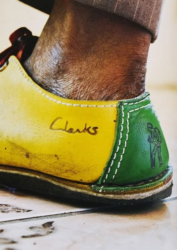 Close-up of a worn yellow and green vintage shoe with the name 'Clarks' written on it, worn by a person with dark skin and pants above the ankle.