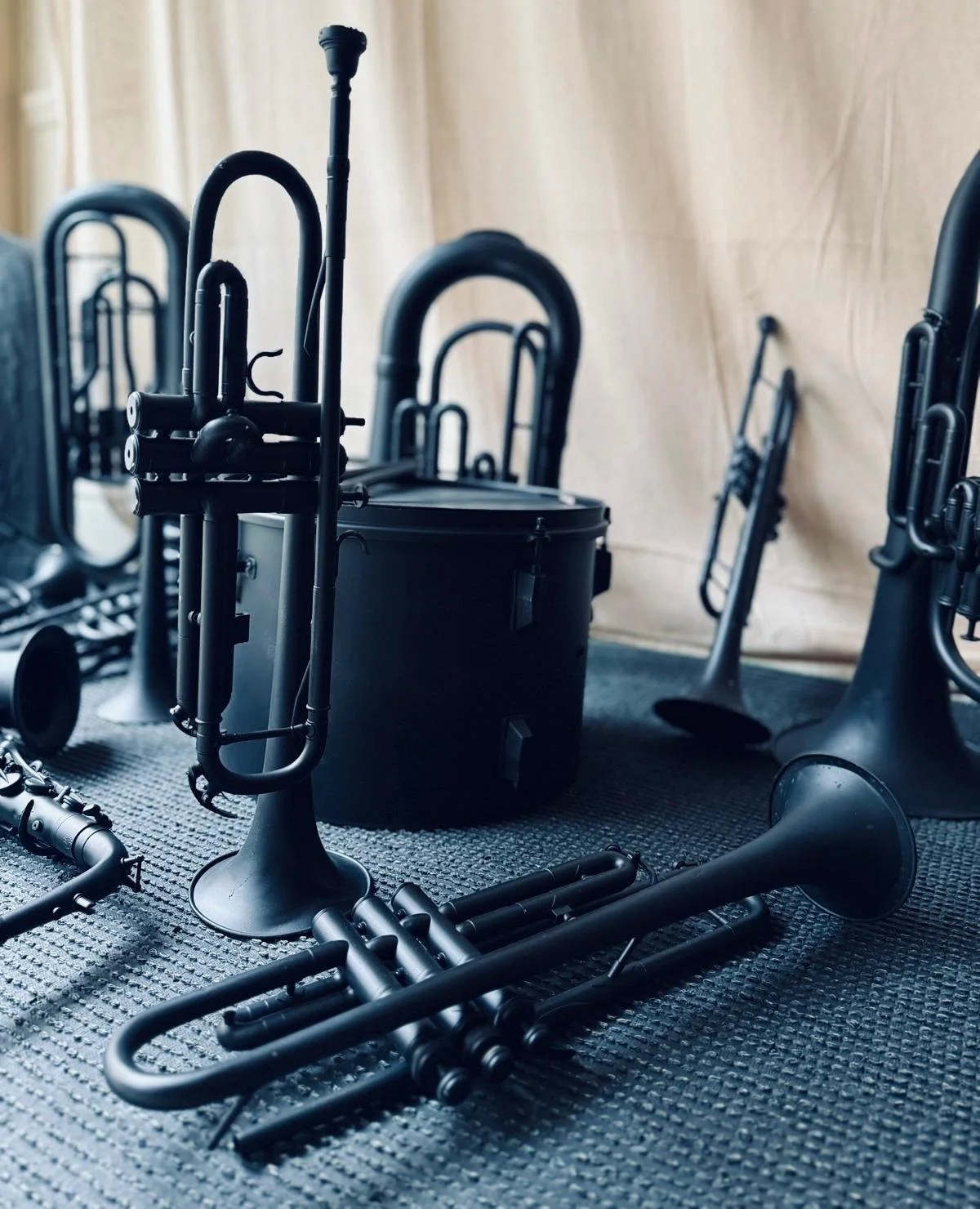 Several black musical instruments including trumpets and a trombone arranged on a dark textured floor near a light-colored wall.