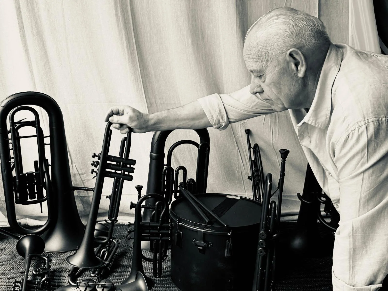 An elderly man with white hair in a light-colored shirt leaning over a black case, reaching towards a group of musical instruments including trumpets, trombones, and a tuba, which are arranged on the floor.