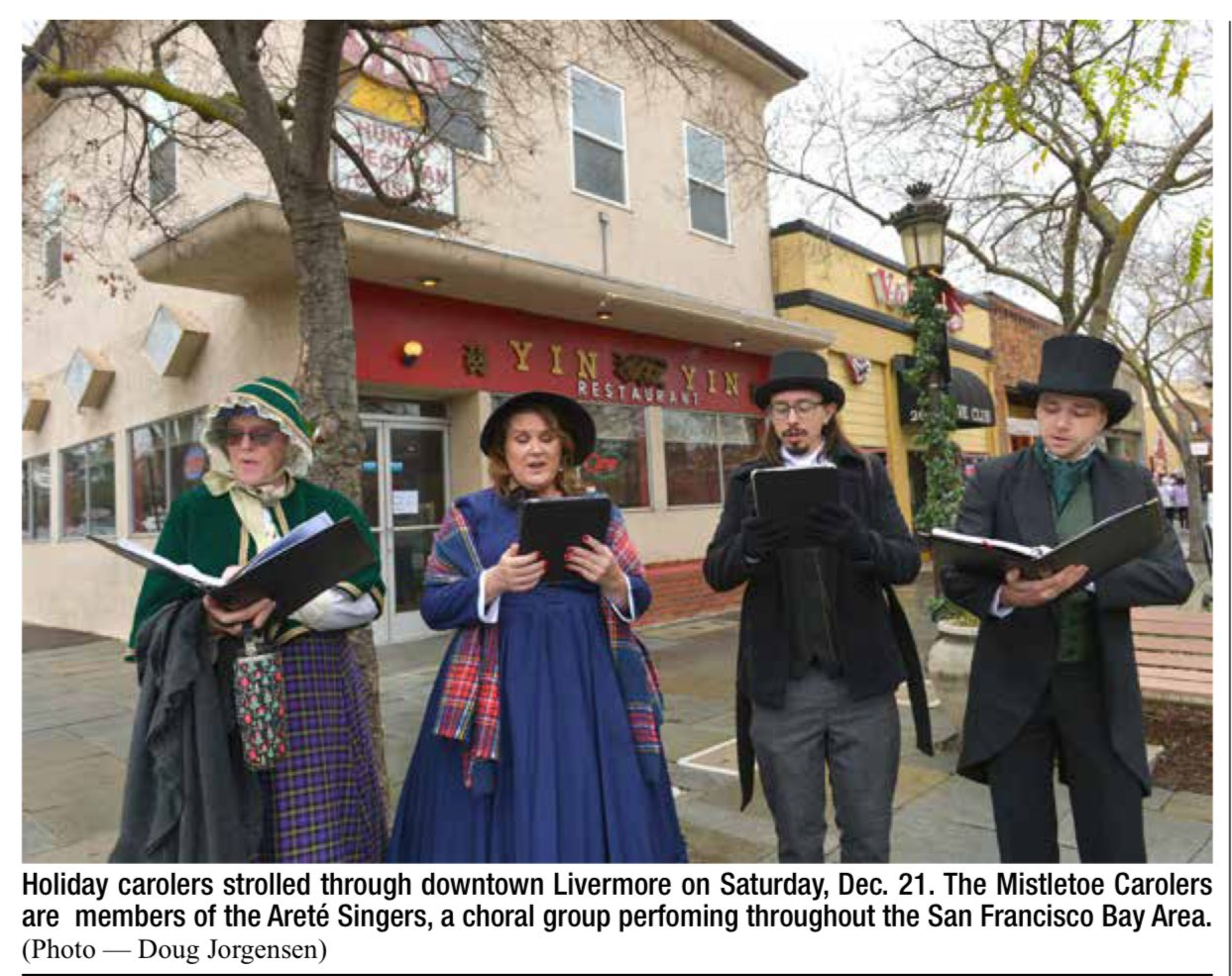 Four people dressed as 19th-century holiday carolers standing on a sidewalk in downtown Livermore, singing from songbooks. Two women and two men, wearing period costumes with hats and coats, are standing in front of a restaurant named 'Yin Yin' and a