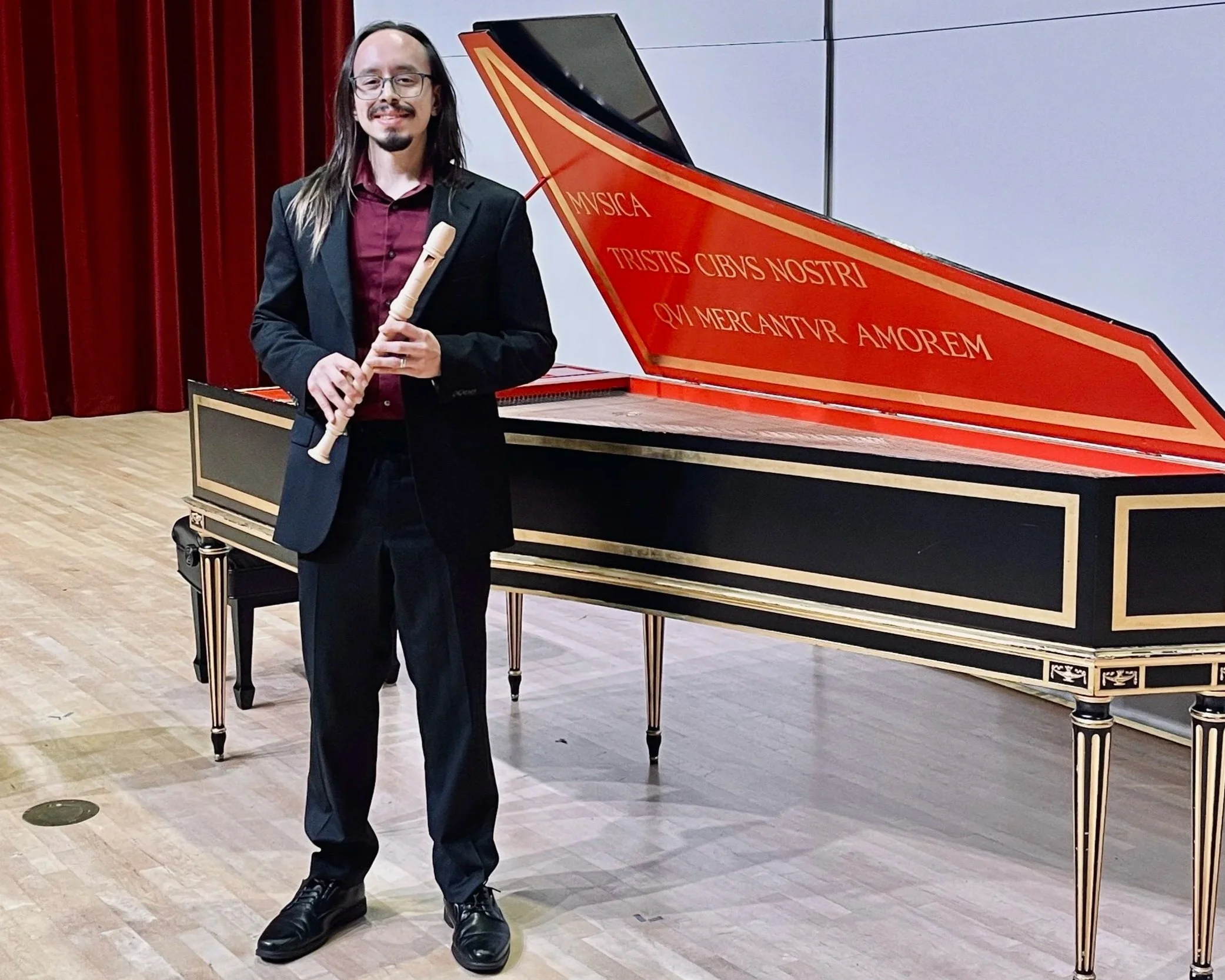 A man with long hair, glasses, in a black suit and maroon shirt holding a recorder, standing next to a red and black piano with Latin inscriptions.
