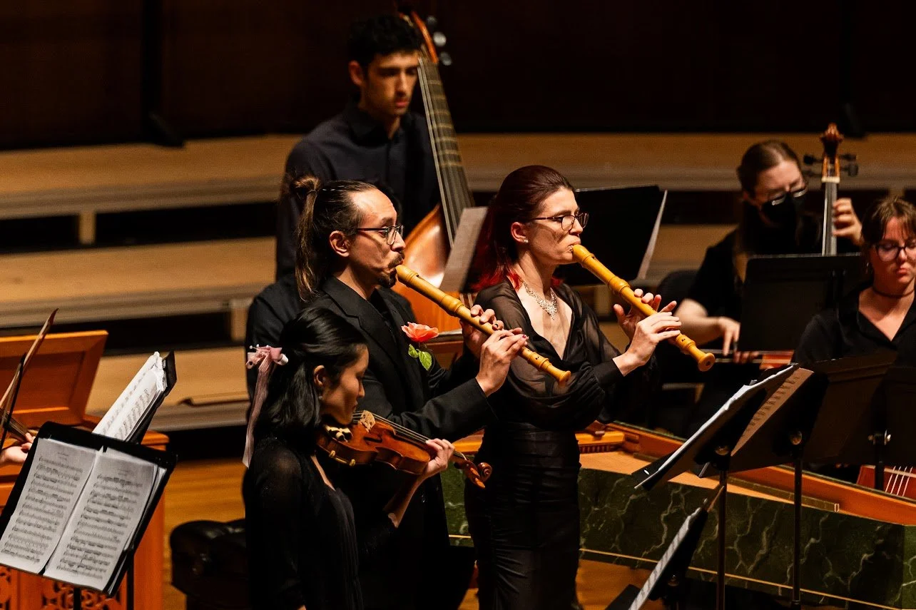 Orchestra performers playing musical instruments during a concert, including two women playing recorders, a woman playing violin, and other musicians in the background.