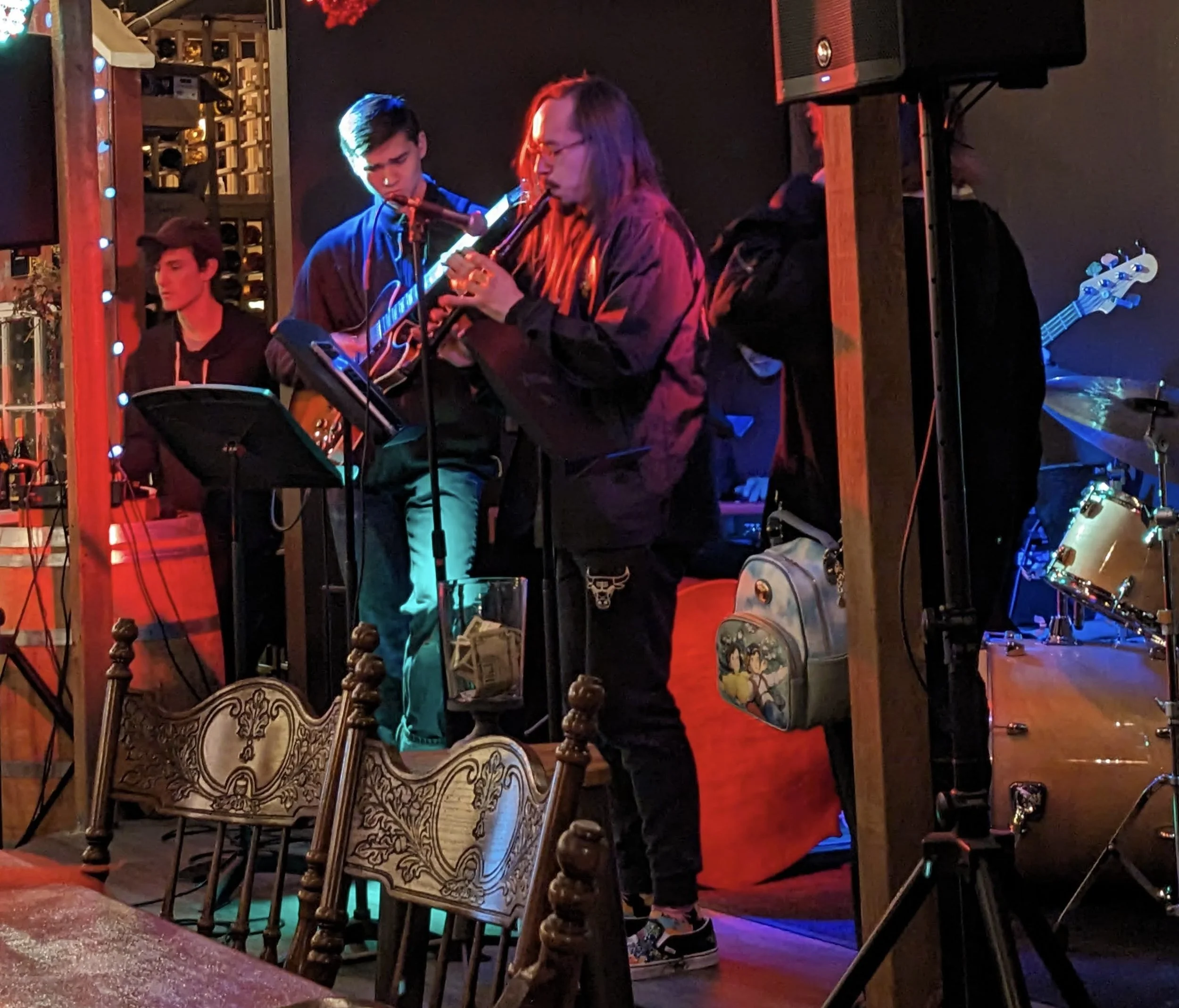 Three young men performing as musicians on a small stage with wooden chairs and a guitar. One is playing a flute, another with headphones holding a bass guitar, and the third is sitting, seemingly waiting or observing. The setting appears to be a coz