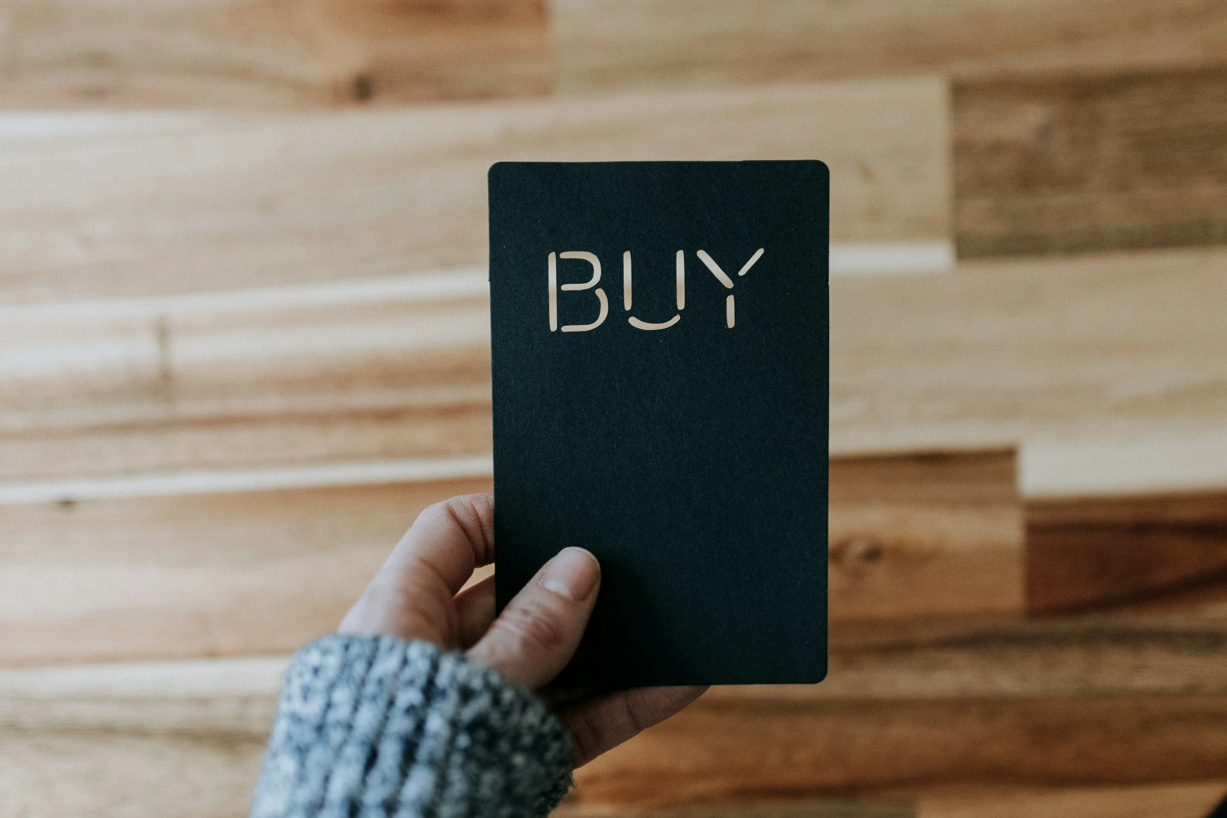 Person holding a black card with the word 'BUY' cut out, against a wooden background.