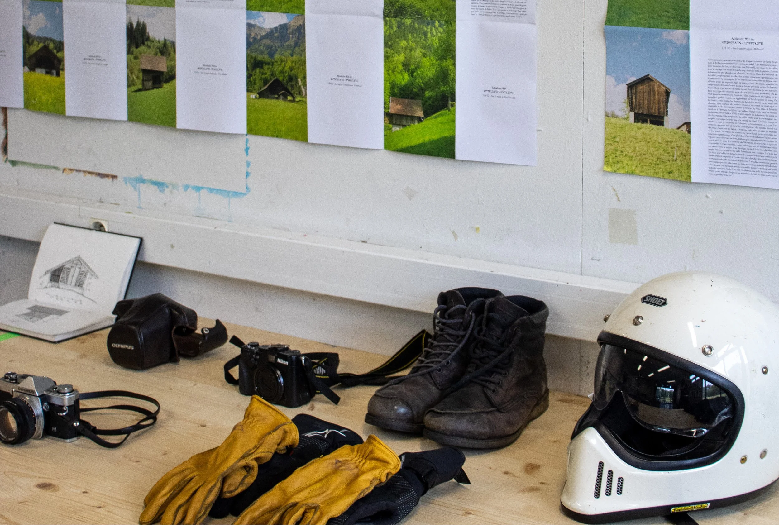 Équipements de moto tout-terrain posés sur un bureau en bois, comprenant un casque, des gants, des bottes, deux appareils photo, une sacoche, un livre avec un croquis de casque en couverture, et des photos de cabanes en montagne affichées sur le mur 