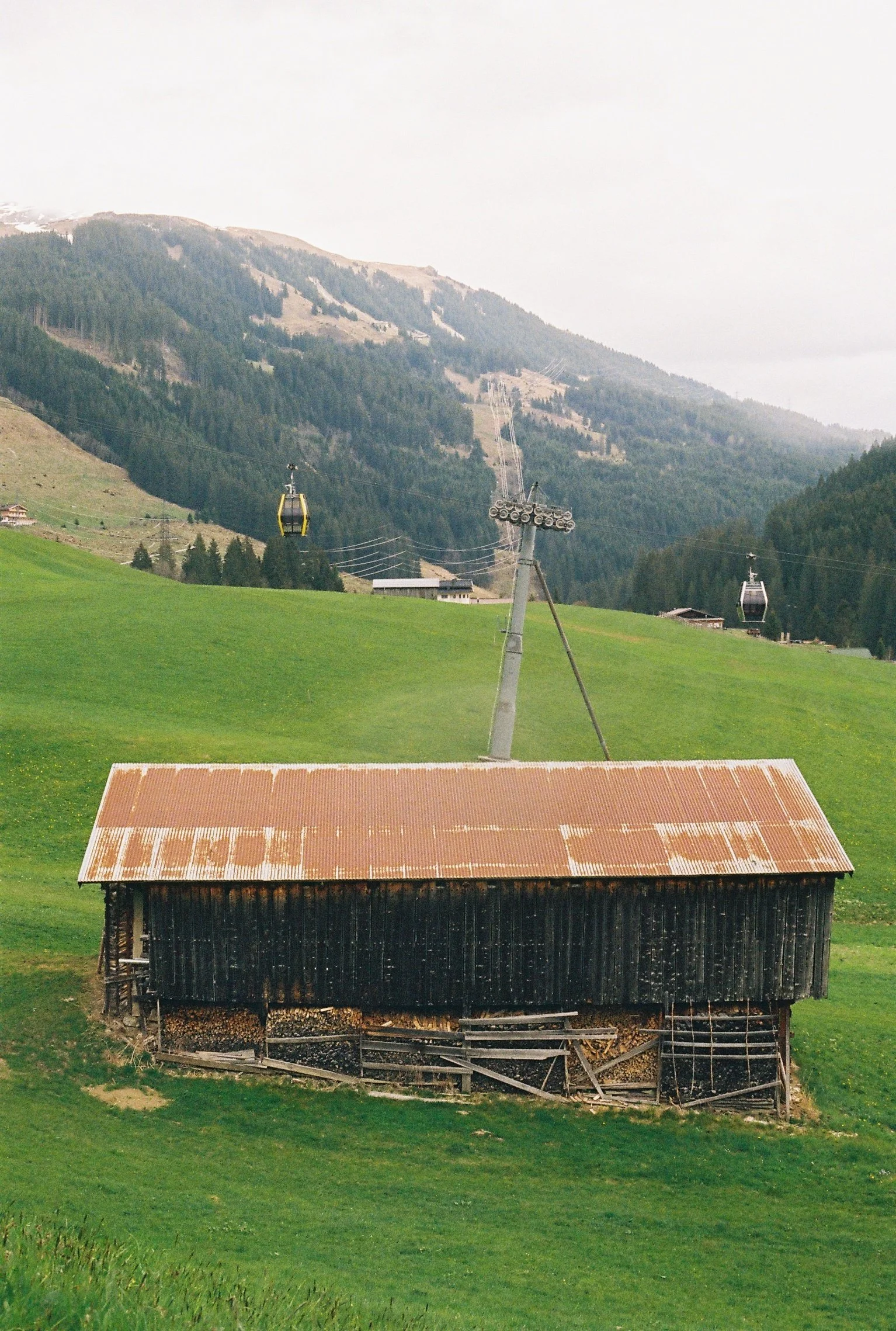 Une vieille cabane en bois avec un toit en tôle rouillée, située sur une prairie verte, avec des montagnes en arrière-plan, et des télécabines suspendues dans le ciel.