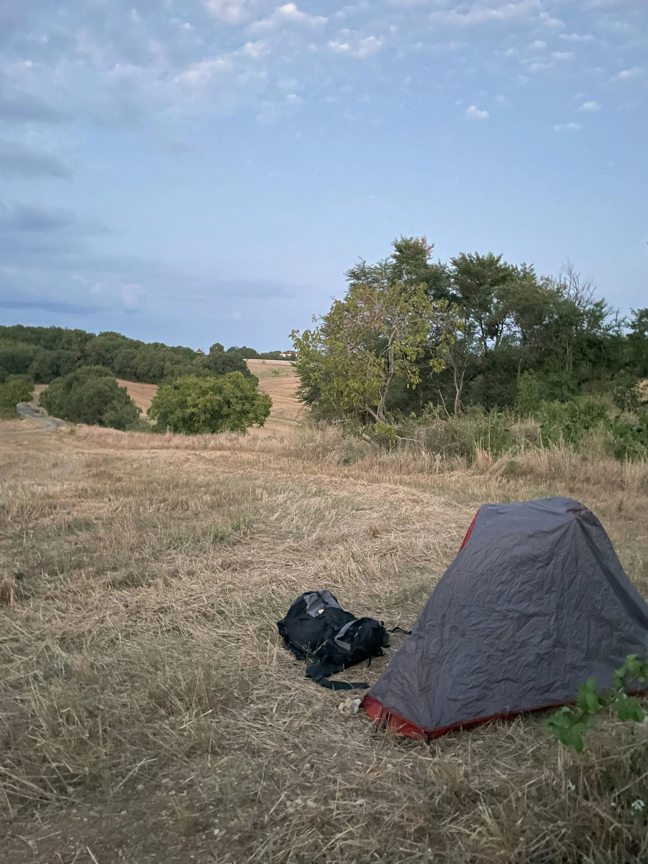 Une tente de camping noire et grise avec un sac à dos noir sur un champ sec en plein air, avec des arbres et un ciel partiellement nuageux en arrière-plan.
