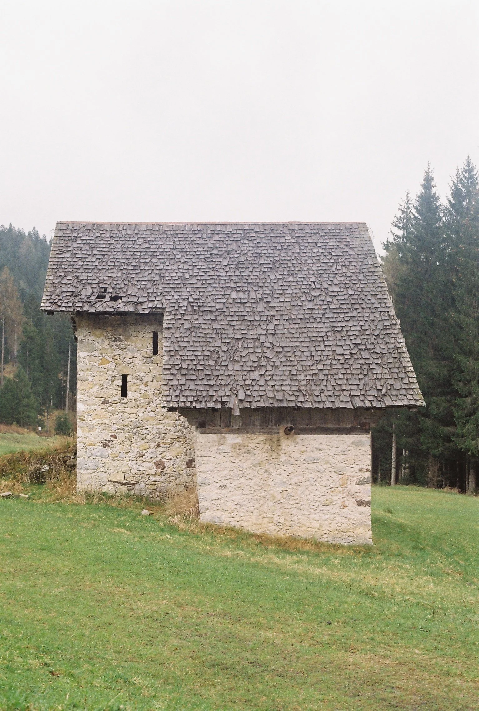Une petite maison en pierre avec un toit en tuiles en schiste, entourée d'herbe et d'arbres en arrière-plan.