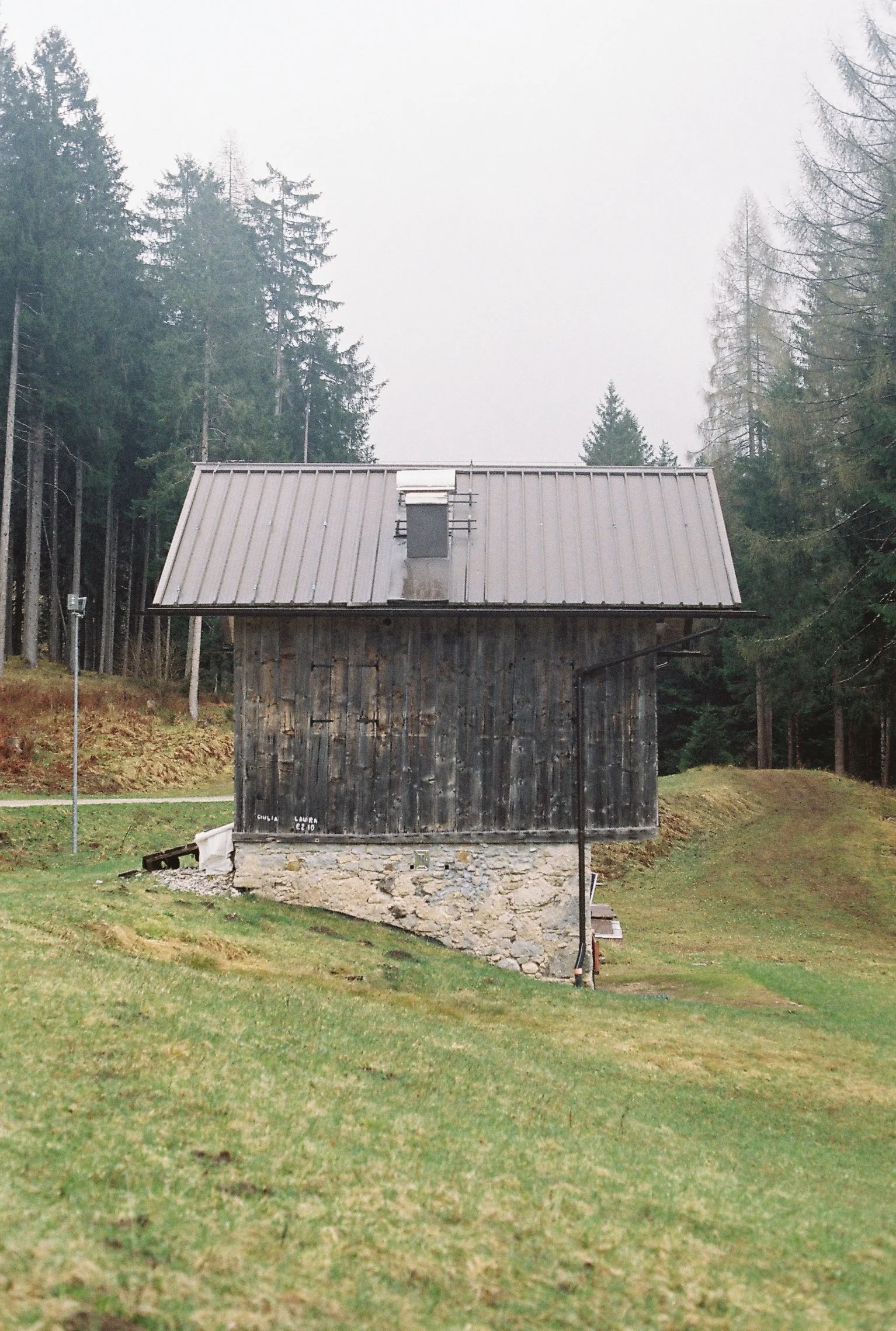 Une cabane en bois sombre sur une fondation en pierre, entourée d'arbres, avec un toit en métal et un petit conduit de cheminée.