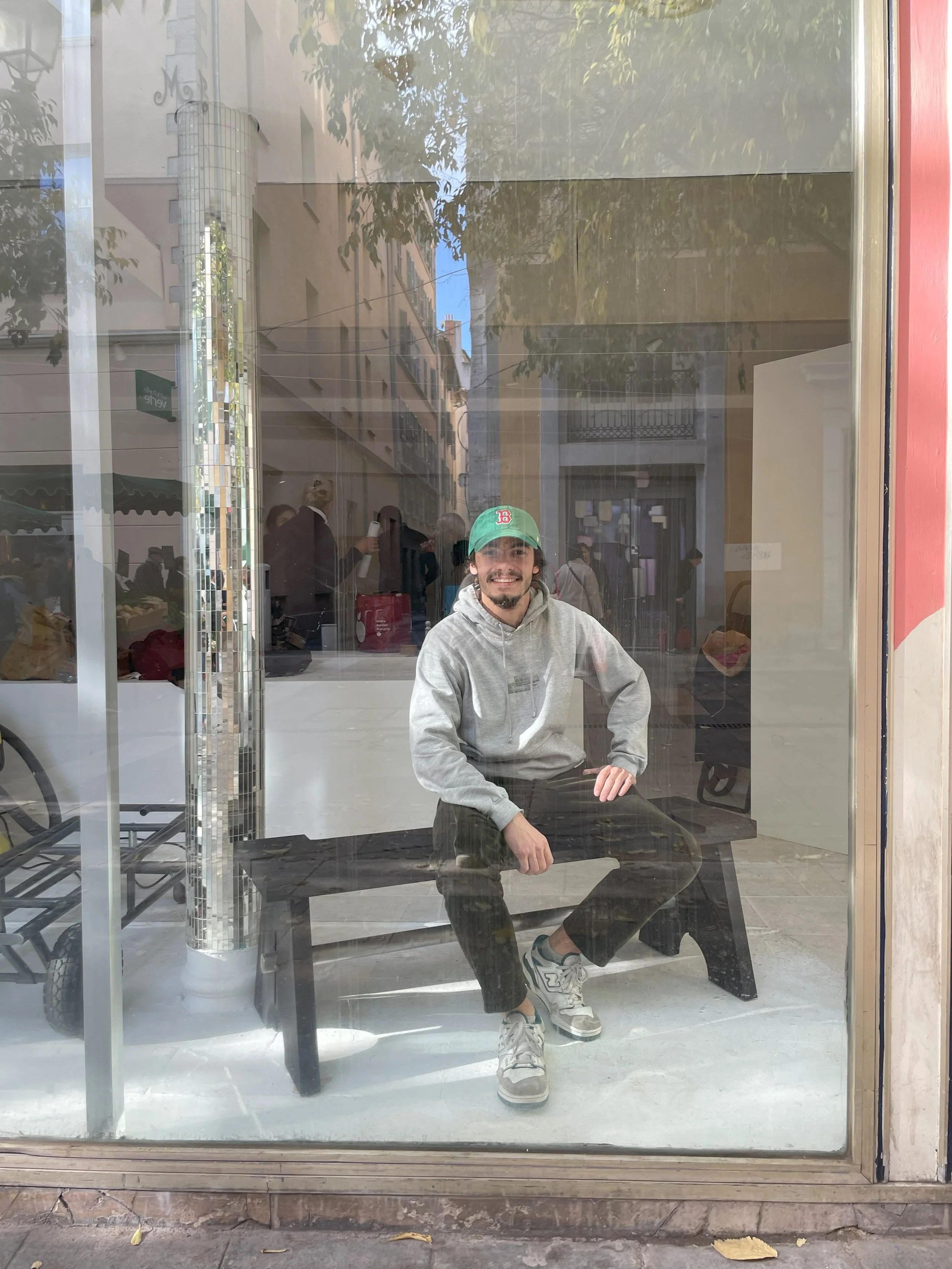 Jeune homme souriant portant un sweat à capuche gris, un pantalon noir, des sneakers grises et une casquette verte, assis sur un banc derrière une vitre d'une boutique.