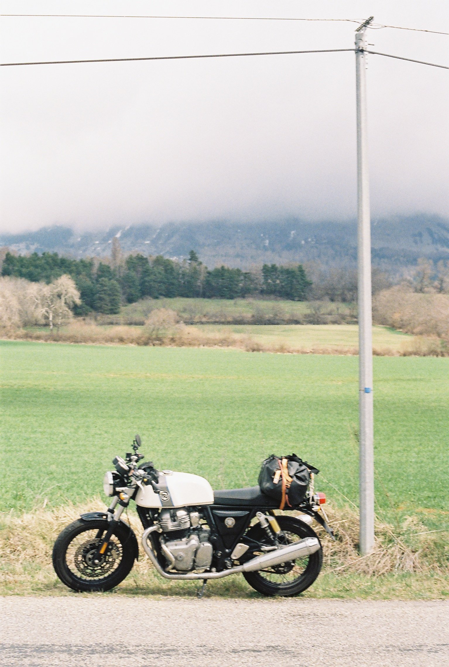 Une moto noire et blanche garée sur le bord de la route à côté d’un poteau électrique dans un paysage rural avec un champ verdoyant et des montagnes en arrière-plan.