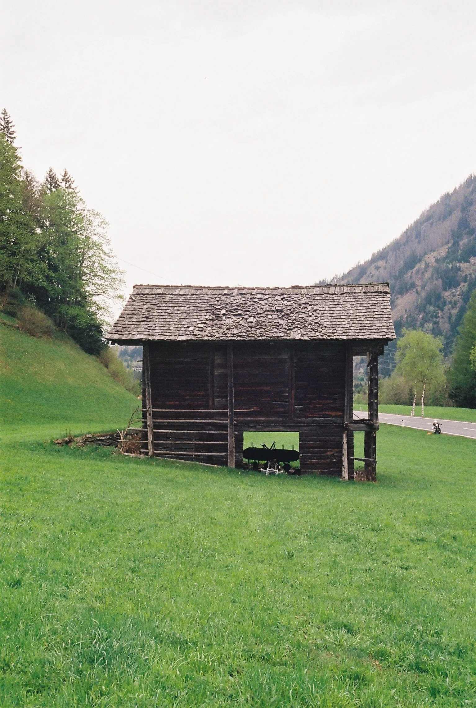 Une vieille cabane en bois sombre située dans un champ vert en zone rurale, avec une route à l'arrière-plan et des montagnes en arrière-plan, sous un ciel nuageux.
