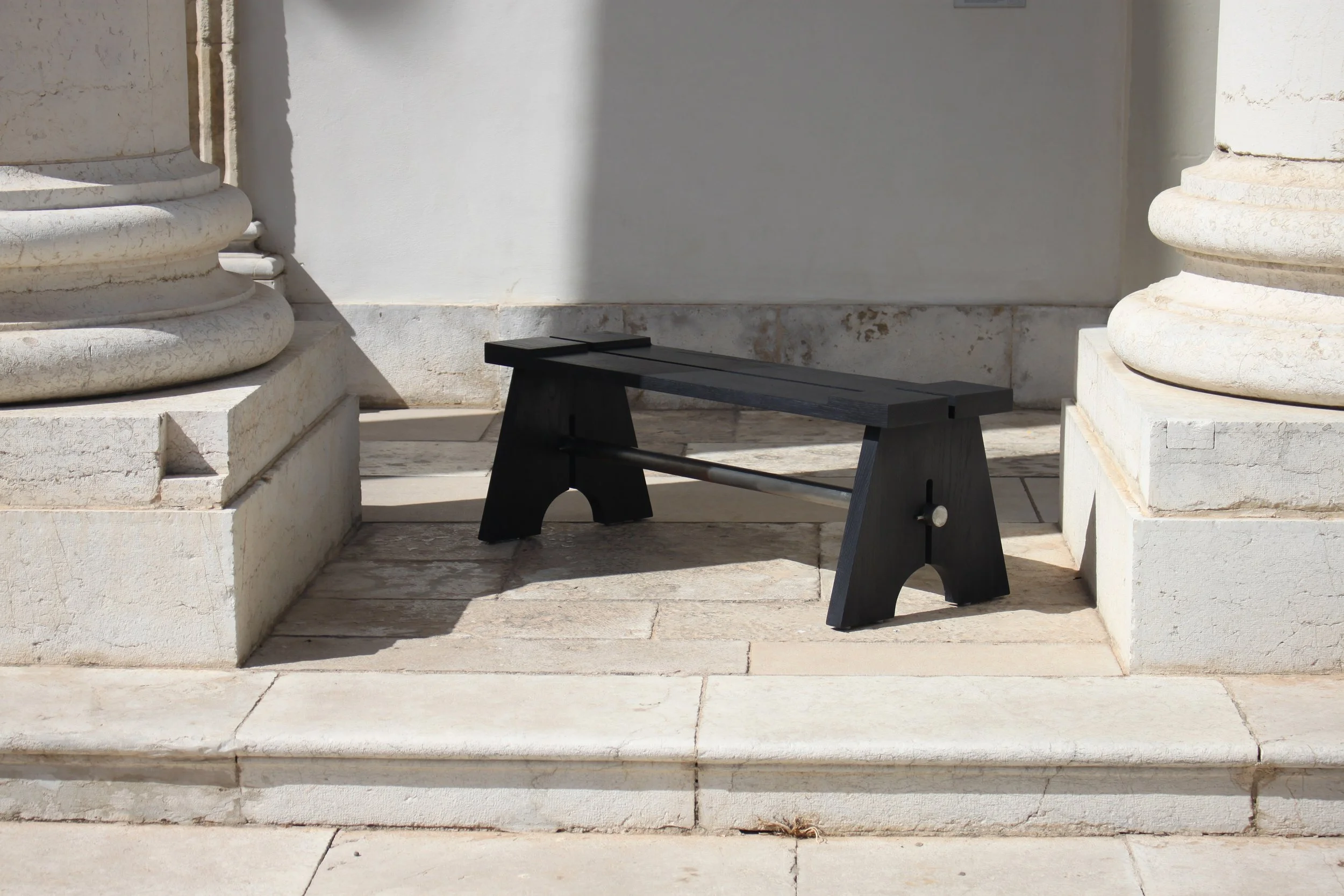Un banc noir en bois placé entre deux colonnes anciennes en pierre sur un sol en pierre blonde.