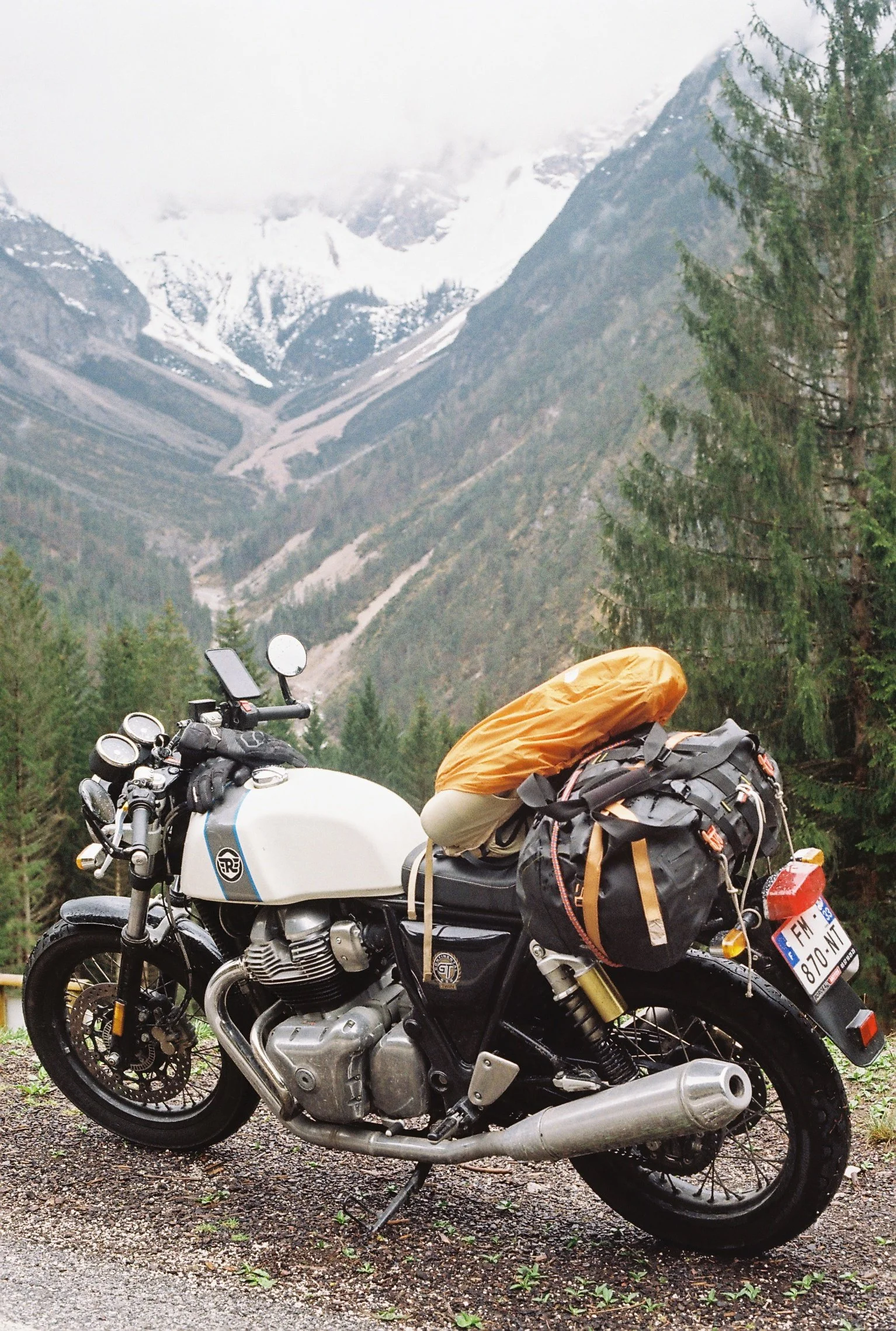 Une moto stationnée dans une vallée alpine avec des montagnes enneigées en arrière-plan, entourée de forêts vertes.