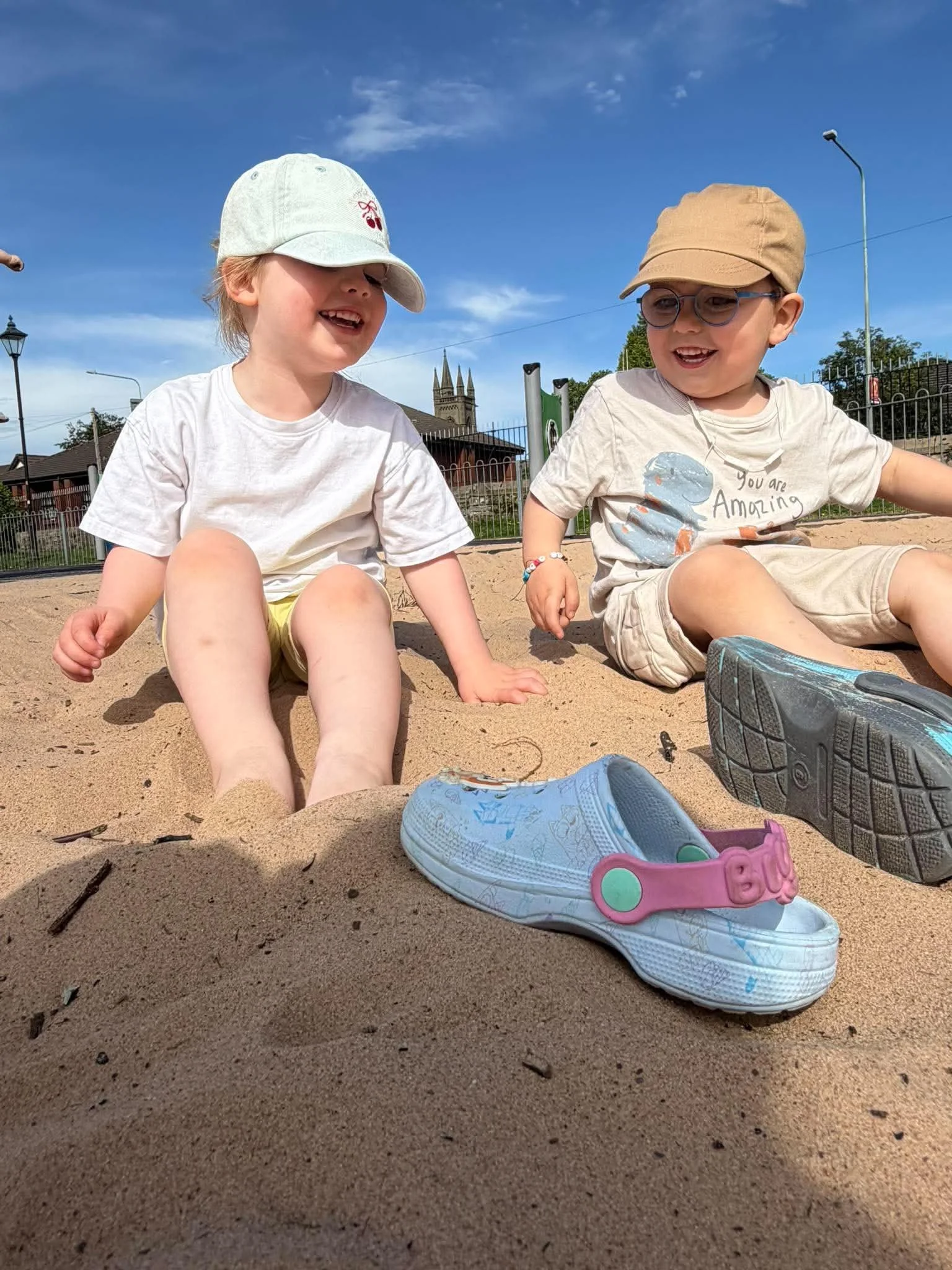 Two young children sitting in a sandbox with shoes in the foreground, smiling and laughing, outdoors on a sunny day with a blue sky, church steeples, and a fence in the background.