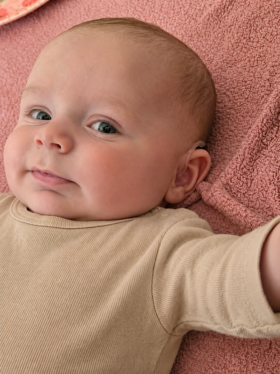 Close-up of a baby lying on a pink textured blanket, wearing a beige shirt, with a slight smile and looking at the camera.