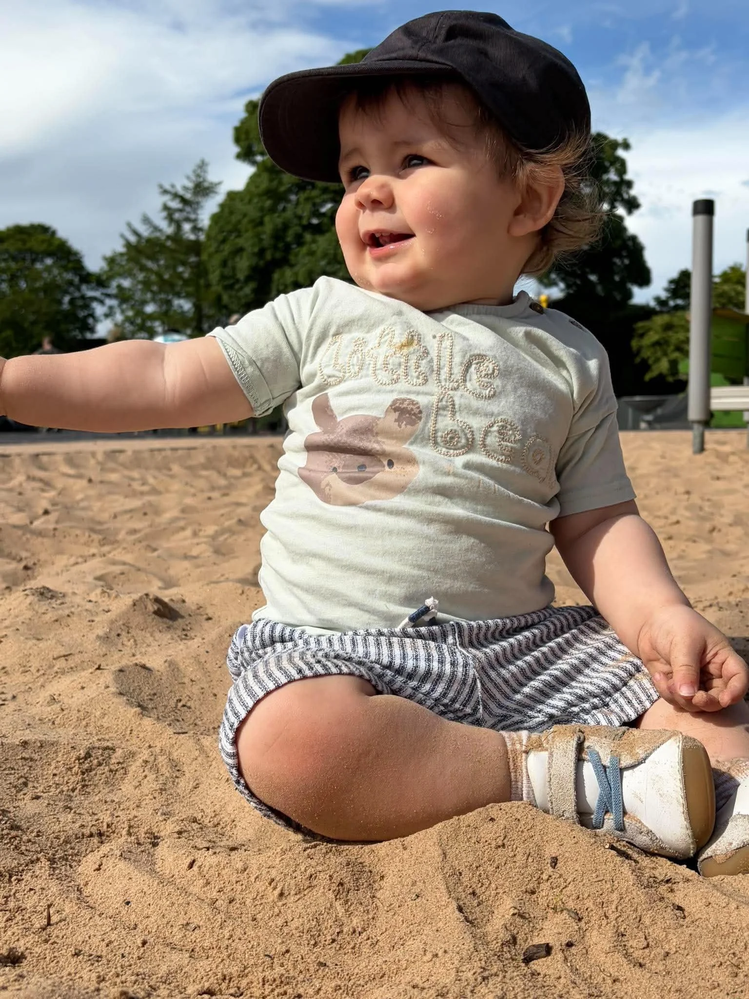 A young child sitting on sandy ground at a park, wearing a baseball cap, striped shorts, a t-shirt with the word 'Chocolate' and a bear graphic, and sneakers. The child has a slight smile and appears to be looking at something in the distance, with trees and a blue sky in the background.