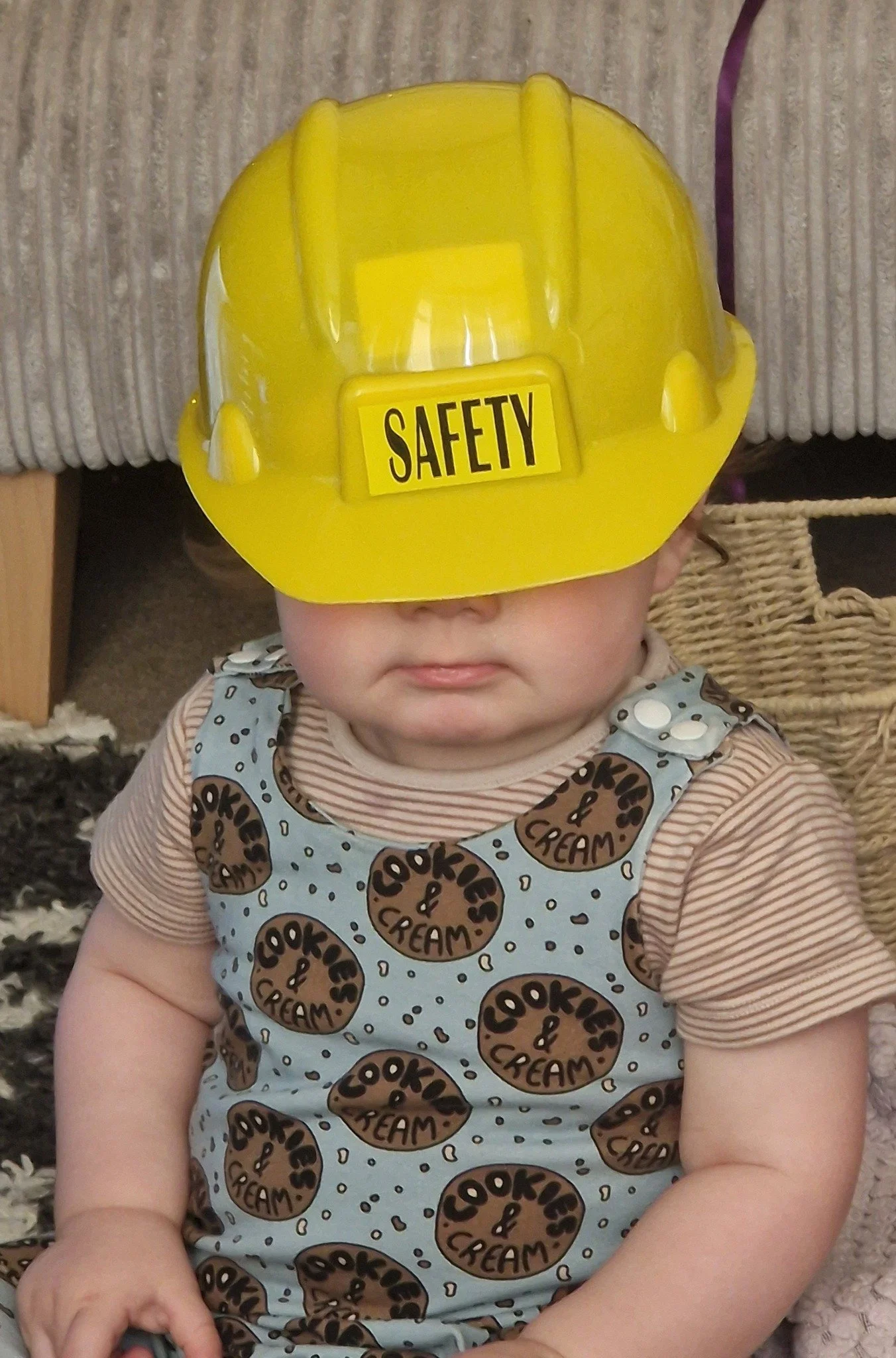 A baby wearing a yellow safety helmet that covers their eyes, sitting on a soft surface with a textured background.