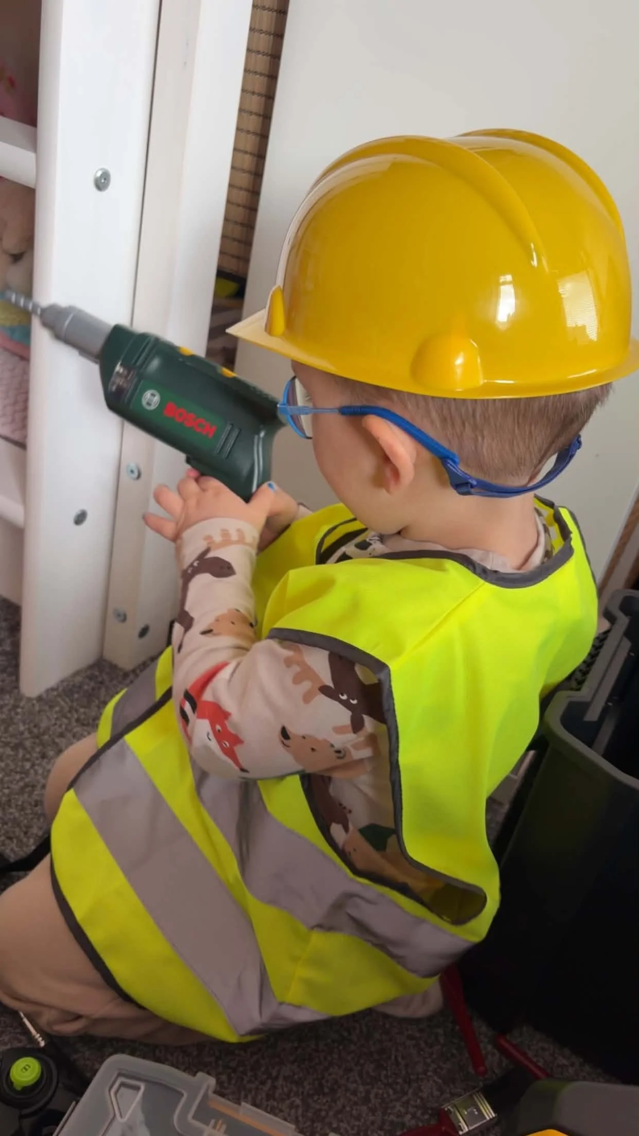 A young boy wearing a yellow construction helmet, glasses, and a neon safety vest is kneeling on the floor and using a cordless drill to screw into a white piece of furniture or paneling.