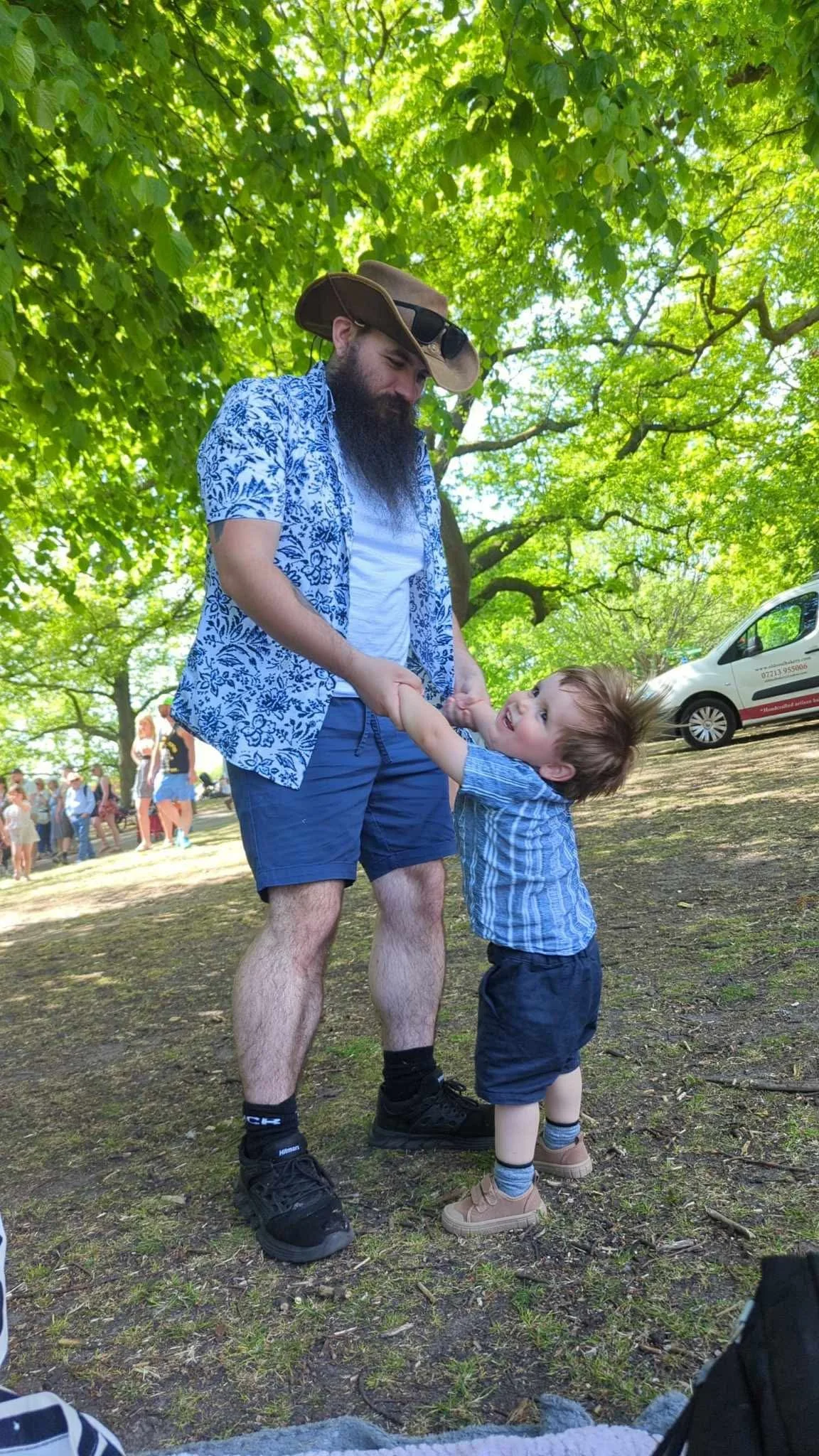 A bearded man in a cowboy hat and sunglasses, wearing a blue floral shirt and shorts, holding the hands of a young boy dressed in a striped shirt and shorts, dancing outdoors under green trees during daytime.