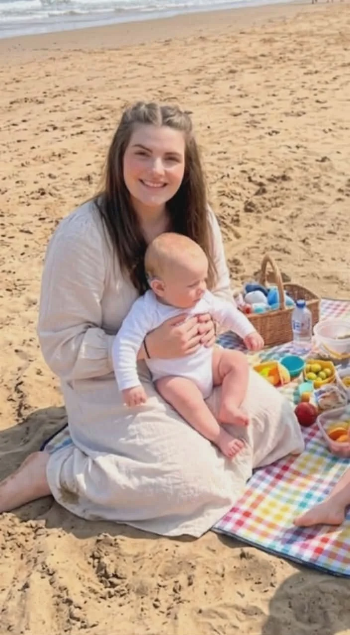 A woman sitting on a blanket on the beach, holding a baby.