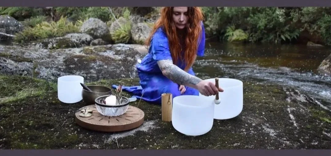 A person with long red hair, tattoos, and a blue dress sits by a river, playing singing bowls placed on a mossy rock.