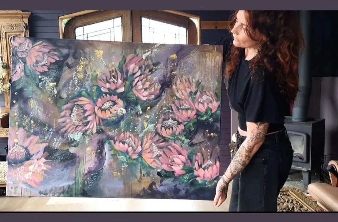 jasmine Hinchliffe artist stands with her large abstract painting of pink flowers with green leaves, gold accents, and splashes of color, in a room with wooden furniture and windows.