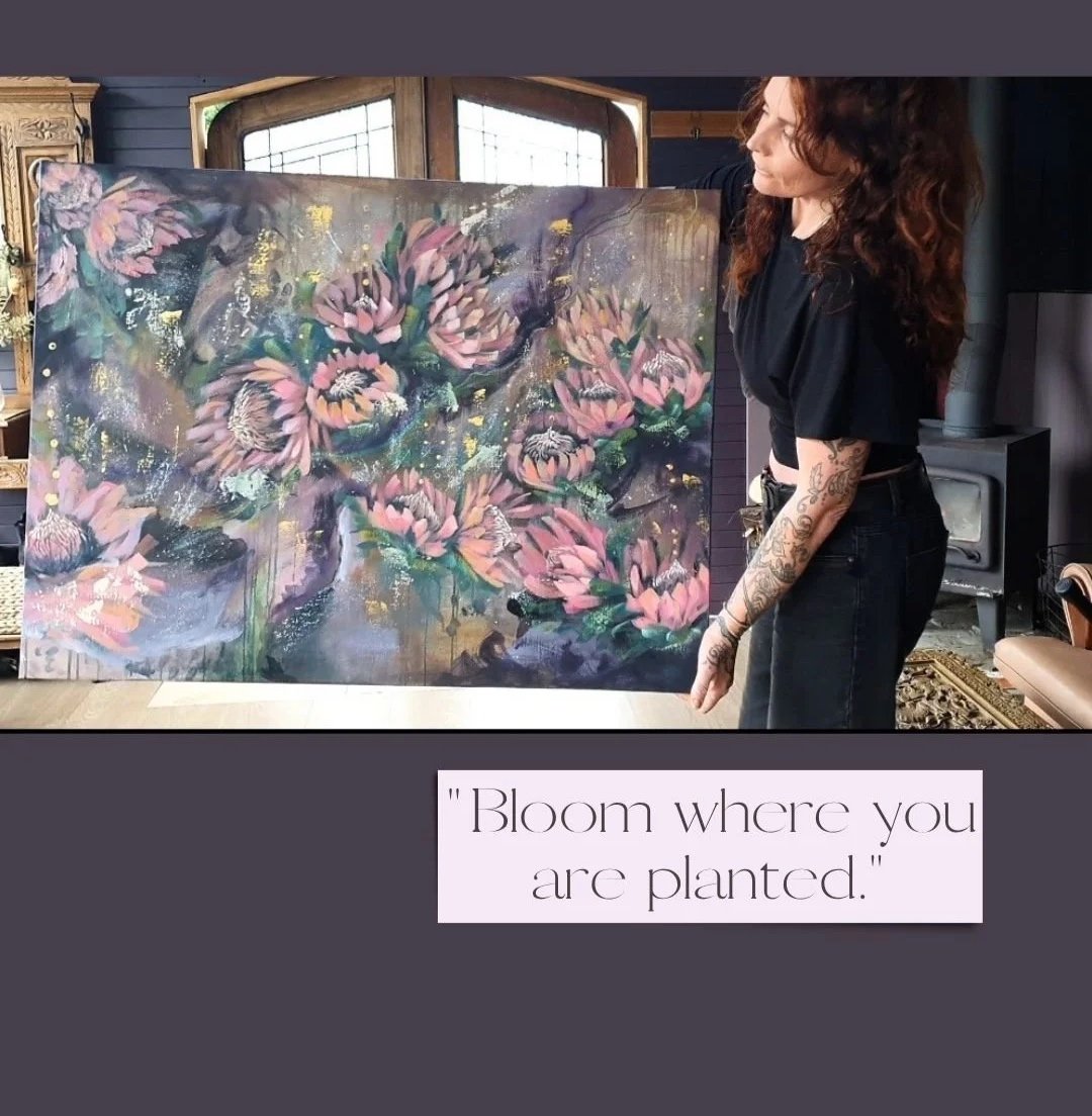 A woman with long curly hair and tattoos on her arm stands next to a large abstract painting of pink flowers with green leaves, gold accents, and splashes of color, in a room with wooden furniture and windows.